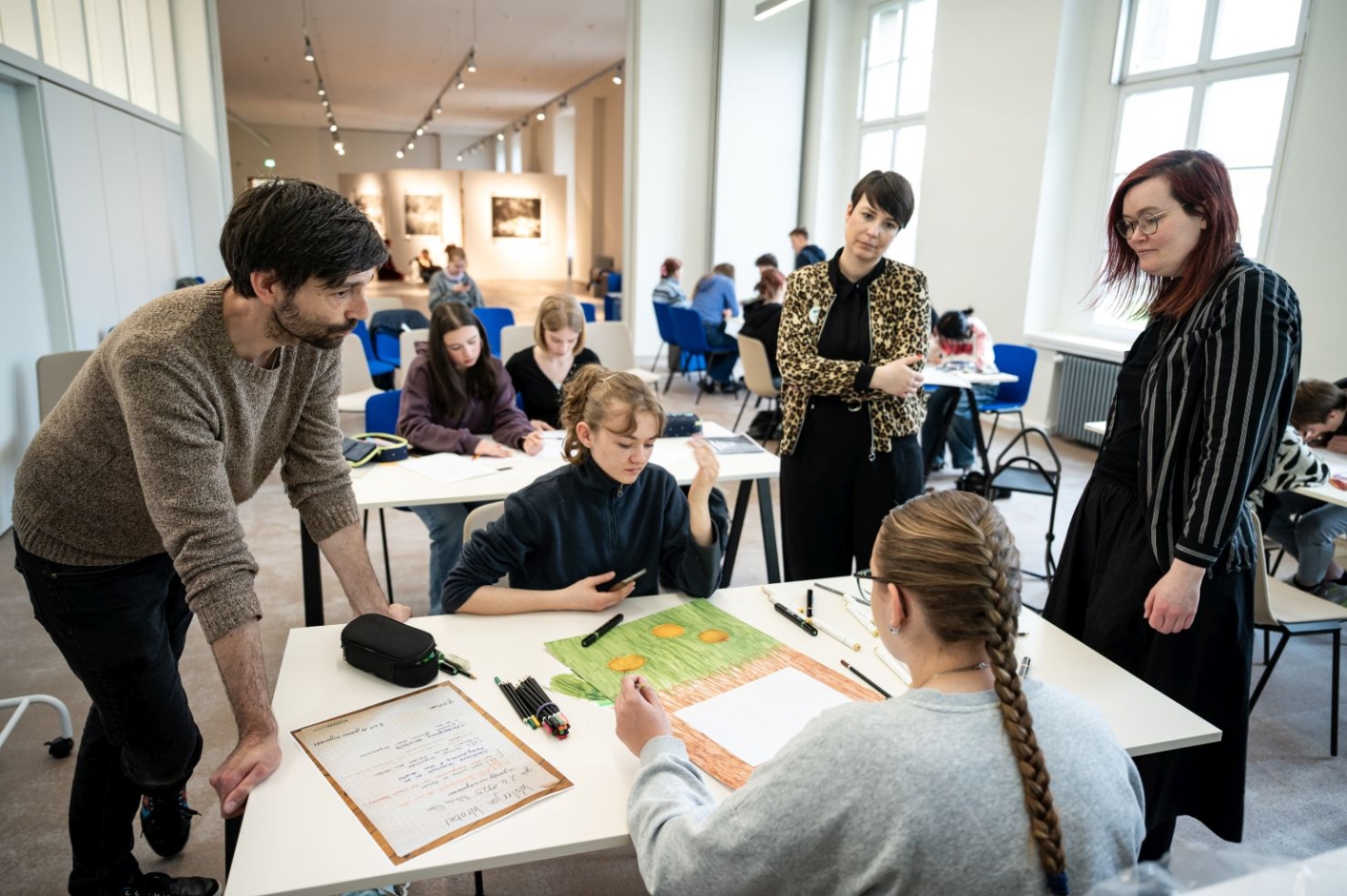 Two schoolgirls are sitting at a table and drawing on a poster. Three people are standing around them to take a look at their work.