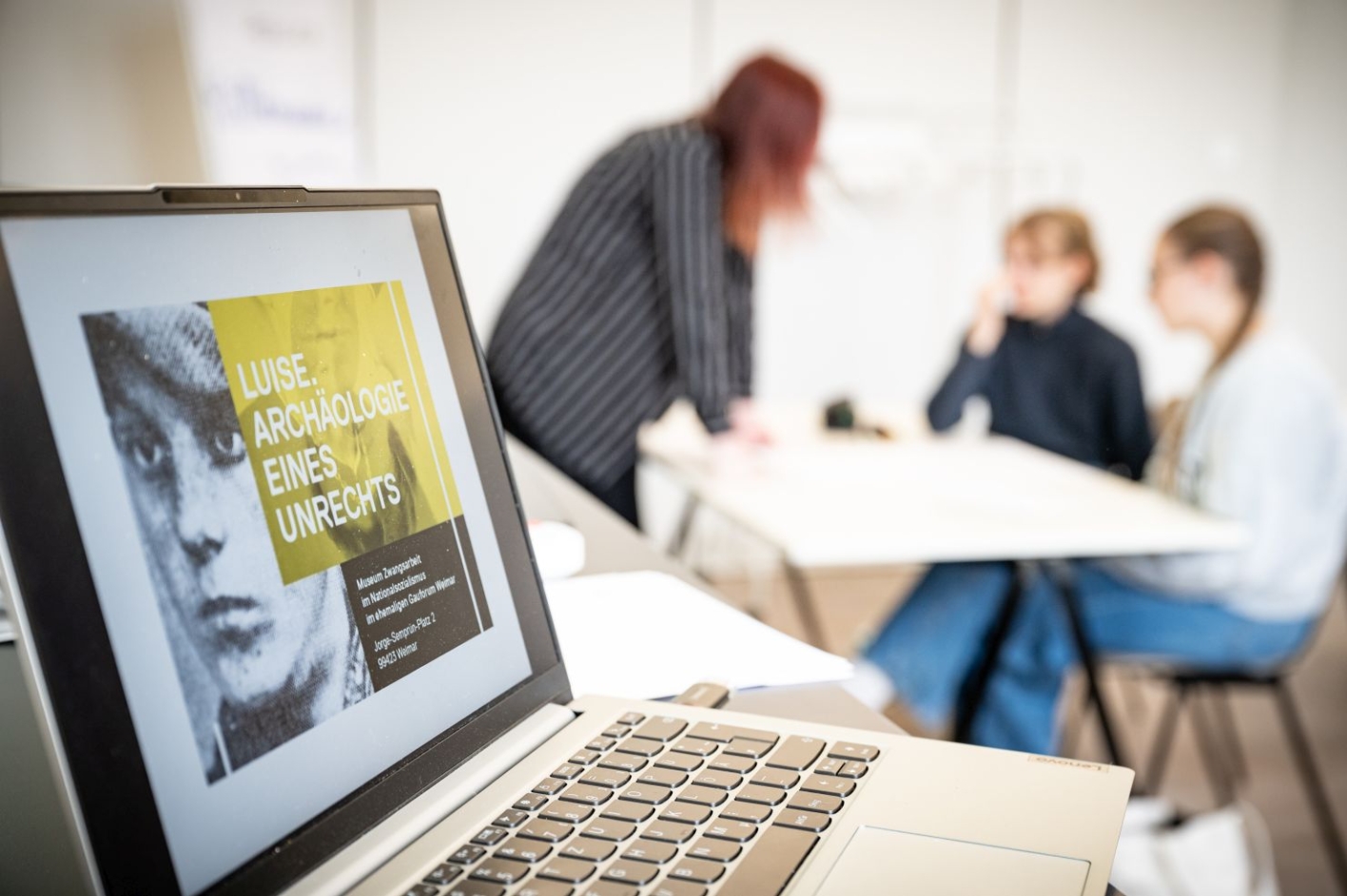 A laptop can be seen in the foreground. This shows the poster advertising the traveling exhibition "Luise. Analogy of an Injustice" was advertised. Three people can be seen in the background. Two are sitting at a table. Another person is standing in front of the table.