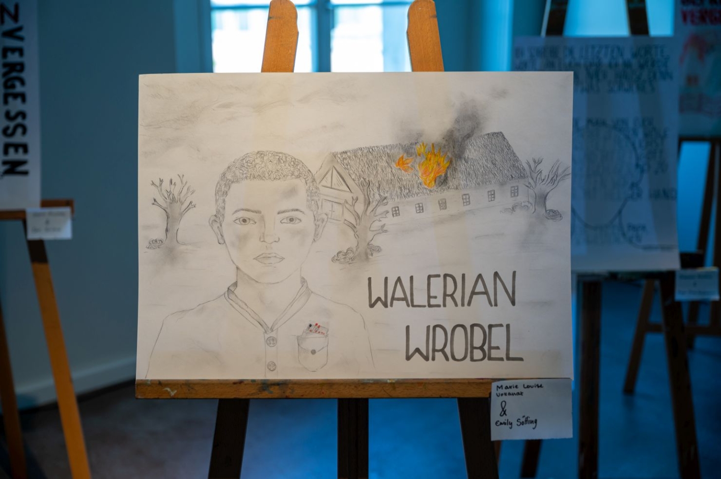 The poster shows the following motif: a sketch of a young person on the left. At the bottom right the lettering “Walerian Wróbel”. A burning house in the background.