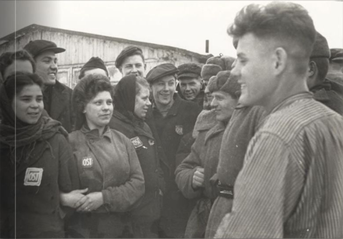Forced labourers with an "Eastern workers' mark" sewn onto their clothing stand opposite Red Army soldiers. Many of the liberated forced labourers are laughing. A barrack can be seen in the background.