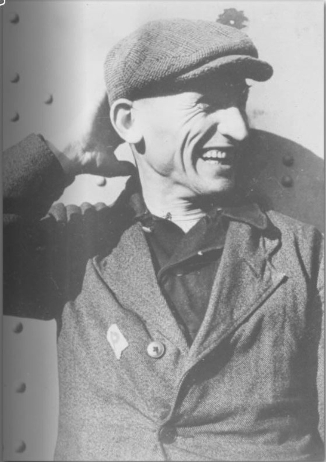 The picture shows Jan Jakubasik. He looks past the camera and laughs. He is holding his right hand behind his head. He is wearing a slouch cap and a jacket with a "Polenabzeichen" sewn onto it.