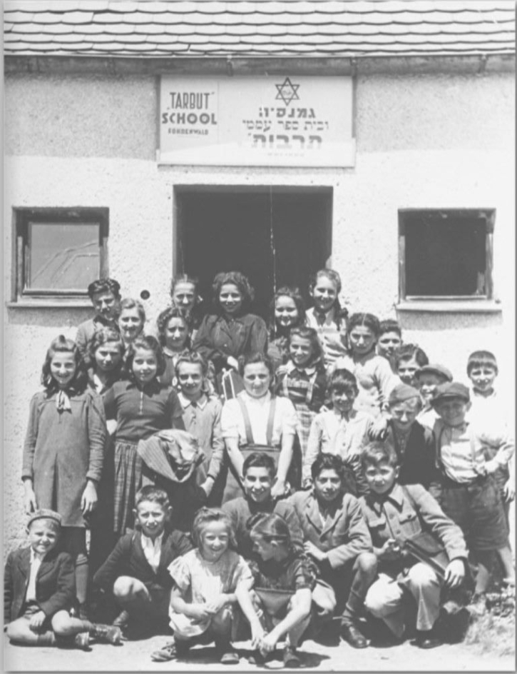 Eine Vielzahl von Jungen und Mädchen unterschiedlichen Alters haben sich vor einem Gebäude versammelt. Über der Gebäudetür befindet sich ein Schild mit der Aufschrift "Taburt School".