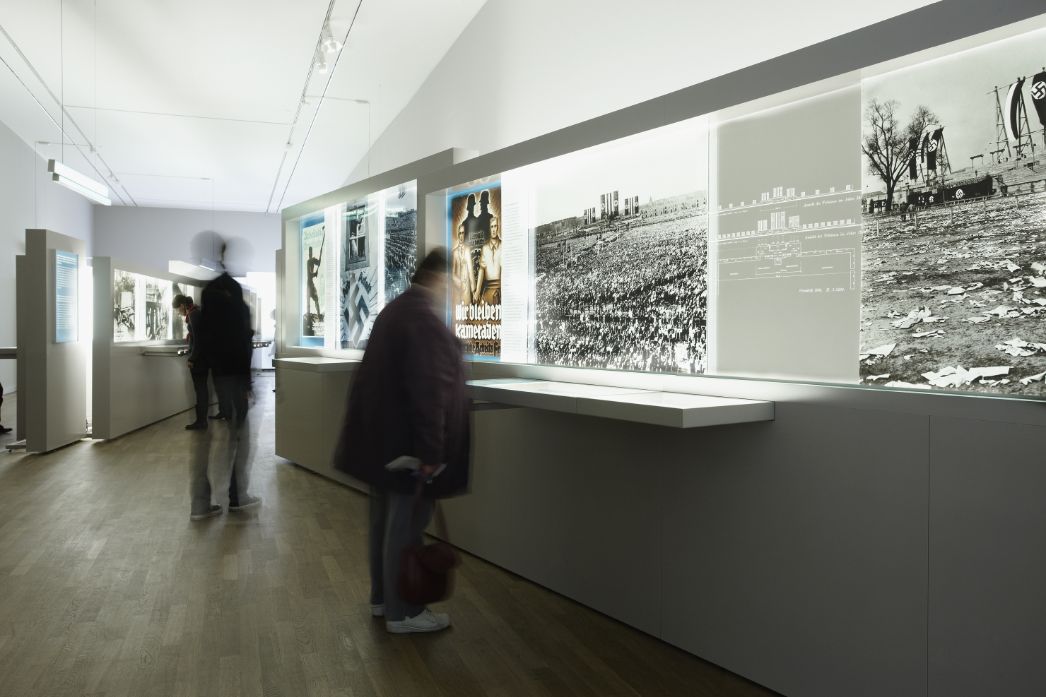 The first part of the exhibition can be seen, which works with large posters and enlarged photographs placed along the walls. The picture shows two people looking at the exhibition walls while moving.