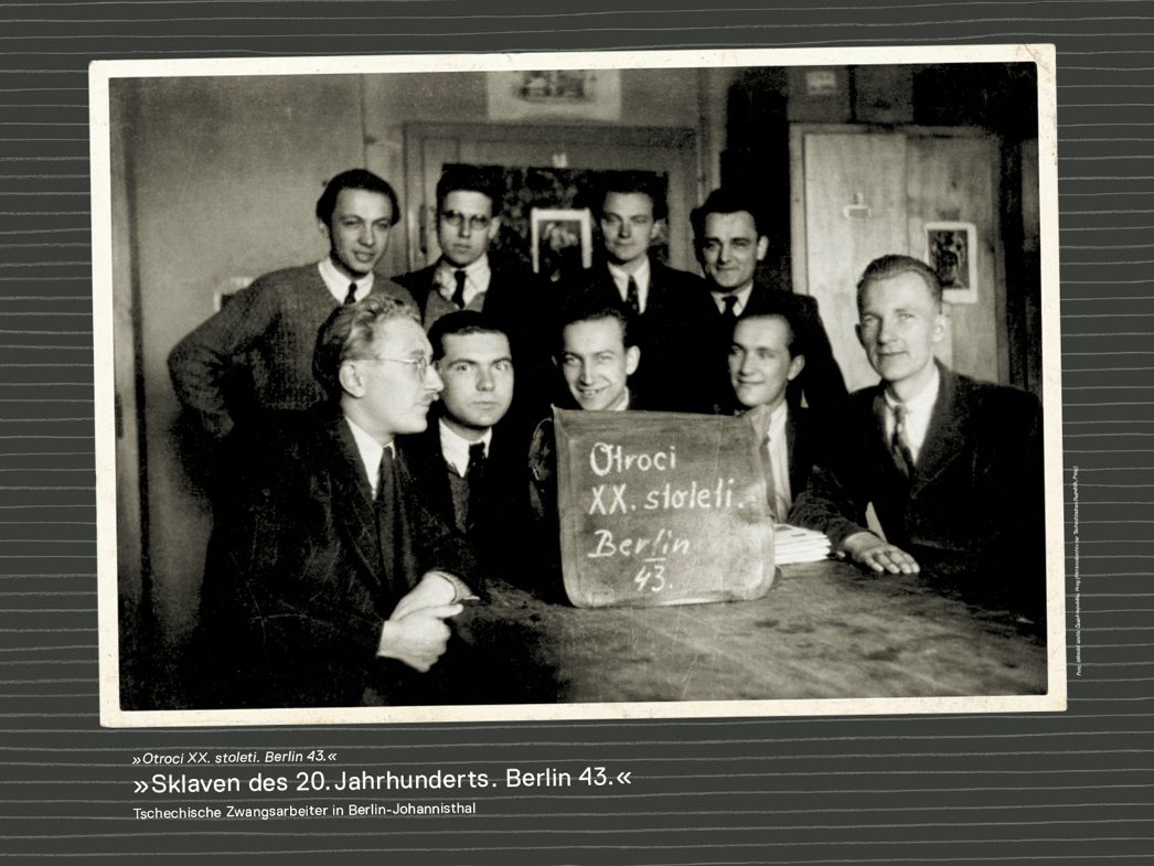 The picture shows 8 men. Four of them are sitting at a table, another four are standing behind them. In front of them on the table is a wooden sign painted with chalk. On it are the words "Slaves of the 20th century. Berlin 43." in Czech