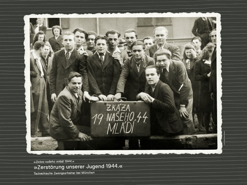 The picture shows a group of young men posing for a photo. In the center of the picture, two of the men are holding a suitcase written on with chalk. It reads "Destruction of our youth 1944" in Czech