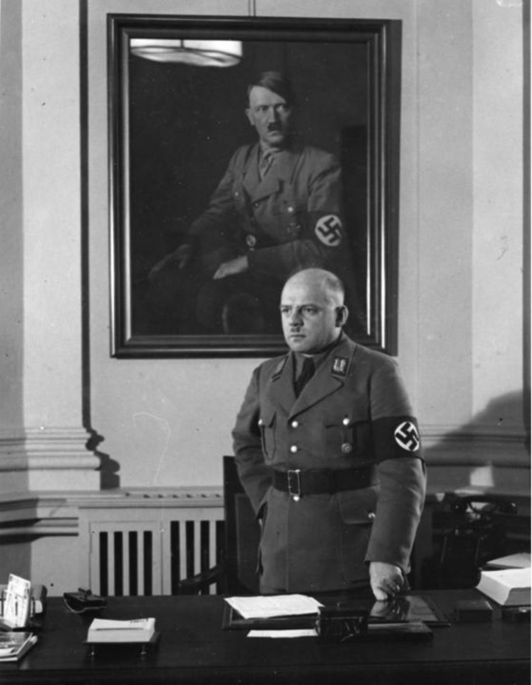 Zu sehen ist ein glatzköpfiger Mann in Uniform, der hinter seinem Schreibtisch, vor einem Portrait von Adolf hitler steht.