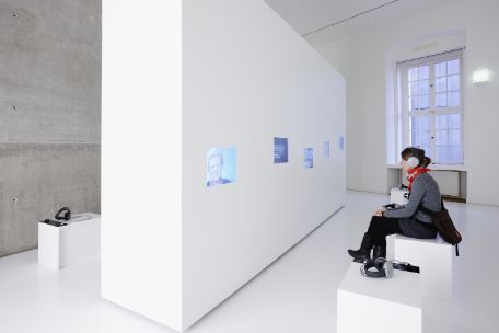 You can see a monochrome white room with a thick white wall. Screens are embedded in this wall. In front of each of these screens is a stool with headphones. On the right side of the picture, a young woman is sitting on one of these stools, wearing the headphones, and looking at the screen in front of her.