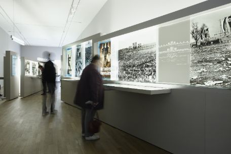 Zu sehen ist der ertse Teil der Ausstellung, welcher mit großen Plakaten und vergrößerten Fotografien arbeitet, die Entlang der Wände angebracht sind. Auf dem Bild sind Zwei Personen zu erkennen, die sich in der Bewegung die Ausstellungswände anschauen.