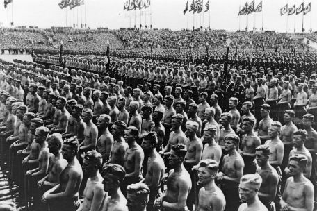 Angehörige des "Reichsarbeitsdienstes" auf dem Parteitag der NSDAP in Nürnberg sitzen in Reih und Glied auf dem Boden. Im Hintergrund sind NS-Flaggen zu erkennen.