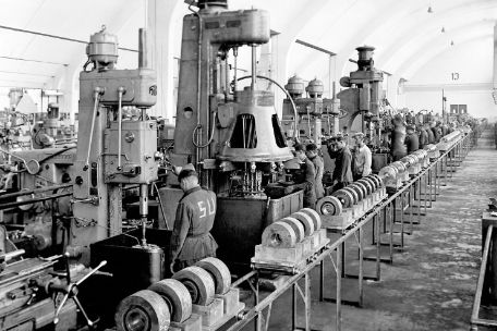 In einer Fabrikhalle arbeiten mehrere Männer. Die Maschinen, an denen sie arbeiten, sind nebeneinander aufgereiht. Auf den Uniformen einiger Männer ist die Aufschrift "SU" zu erkennen.