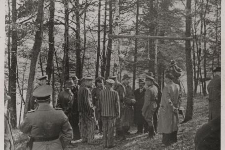Vor einem Galgen in einem Waldgebiet steht eine Männergruppe. Einige der Männer tragen zivile Kleidung, andere NS-Uniformen, wieder andere tragen die gestreifte Kleidung von KZ-Häftlingen.