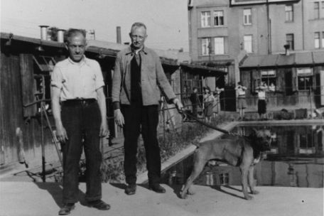 Zwei Männer stehen nebeneinander. Der rechts stehende Mann hält einen Hund an der Leine. Im Hintergrund sind Baracken zu sehen. 