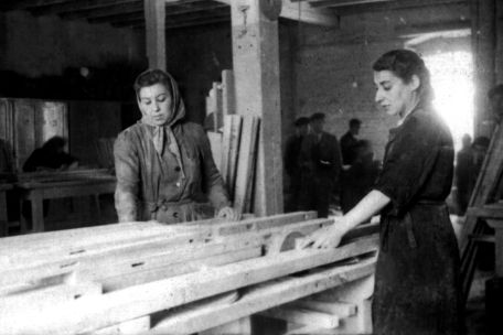 Zwei Frauen verrichten Holzarbeiten in einer Tischlerwerkstatt. Der Zustand ihrer Kleidung ist schlecht.