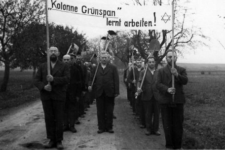 Zu sehen ist eine in Reih und Glied aufmarschierte Kolonne Zwangsarbeite. Die forderen beiden Männer halten ein Trasparent hoch. Es hat die Aufschrift: "Kolonne Grünspan lernt arbeiten!" Daneben ist ein Davidstern aufgemalt.