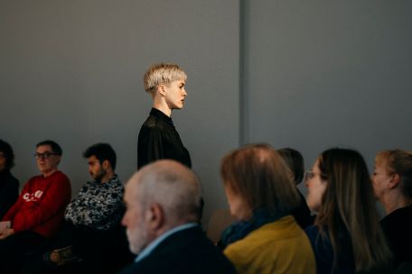 Eine Person mit kurzem, hellem Haar steht aufrecht und ruhig zwischen sitzenden Zuschauer*innen. Ihr seitliches Profil wirkt konzentriert und gesammelt, während das Publikum um sie herum beobachtet. Die Szene hat etwas Performatives: Die klare Haltung der stehenden Figur setzt einen bewussten Kontrast zur sitzenden Menge und lenkt die Aufmerksamkeit auf ihre Präsenz im Raum.