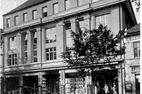 Schwarz-Weiß-Foto eines alten Gebäudes mit dem Schild "KAUFHAUS" und einer gepflasterten Straße im Vordergrund.