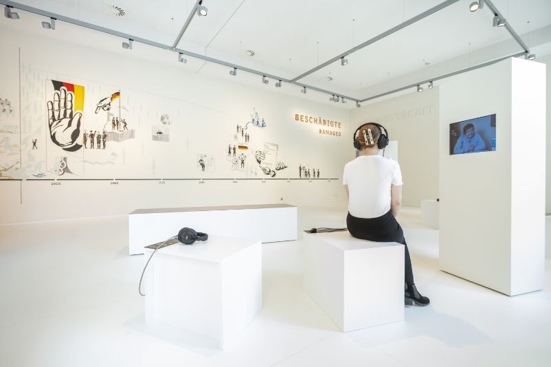 You can see a monochrome white room with a thick white wall. Screens are embedded in this wall. In front of each of these screens is a stool with headphones. On the right side of the picture, a young woman is sitting on one of these stools, wearing the headphones, and looking at the screen in front of her.