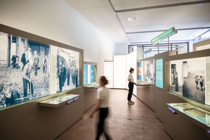 The picture shows two blurred figures standing in front of an exhibition showcase. In the foreground on the left is an exhibition case with a sculpture. It shows figures holding up a swastika on which an eagle is landing.