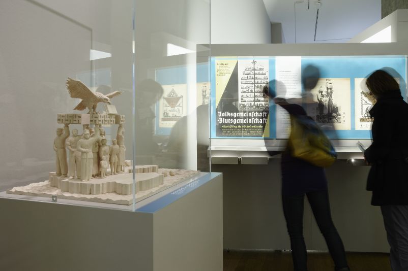 The picture shows two blurred figures standing in front of an exhibition showcase. In the foreground on the left is an exhibition case with a sculpture. It shows figures holding up a swastika on which an eagle is landing.