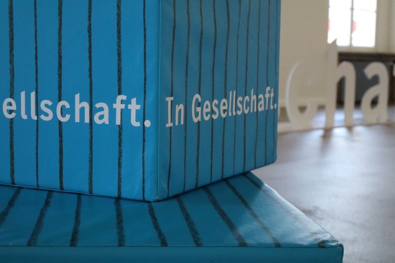 Ein blaues, rechteckiges Sitzpolster steht hochkant im Vordergrund auf der gesamten linken Bildseite. Im Hintergrund bilden weiße Buchstaben, die auf dem Boden vor einem Fenster stehen, das Wort Gesellschfaft.
