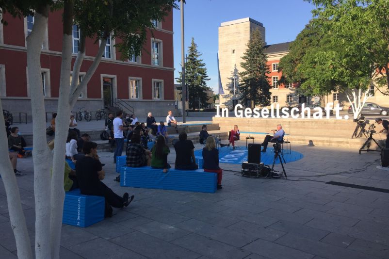 Zu sehen ist der Hesselplatz in Weimar als veranstalltungsort für das Forum "In Gesellschaft", auf dem Blaue blöcke als Sitzmöglichkeiten aufgestellt wurden. Im Hintergrund ist der "Schriftzug: "In Gesellschaft" zu erkennen.