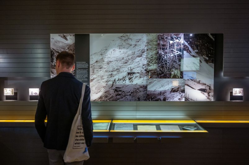 Zu sehen ist ein Mann der vor einer Ausstellungswand steht. Die austellungswand zeigt Fotos von Zwangsarbeitsbaustellen.