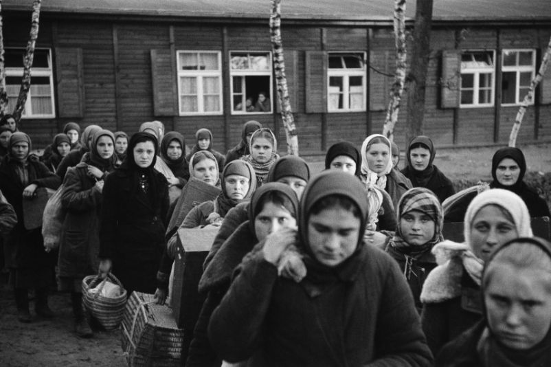 Auf dem Bild ist eine Kolone sowjetischer Zwangsarbeiterinnen zu sehen die vor einer Baracke in Richtung des Fotografen laufen. Ihre Gesichter blicken niedergeschlagen in die Kamera. Sie tragen leichtes Reisegepäck bei sich.