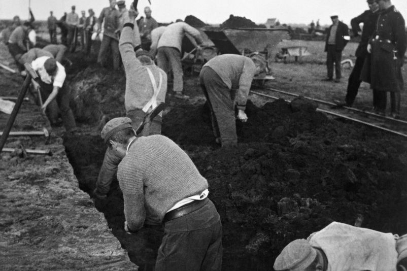 Zu sehen sind Männer die mit Handwerkzeugen Torf stechen. Sie werden von einem Uniformierten Mann am rechten Bildrand beaufsichtigt.