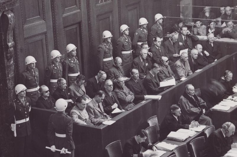 Das Bild zeigt die Anklagten während des Nünrberger Prozesses. Die Anklagebank, auf der sie sitzen, wird von Soldaten bewacht.