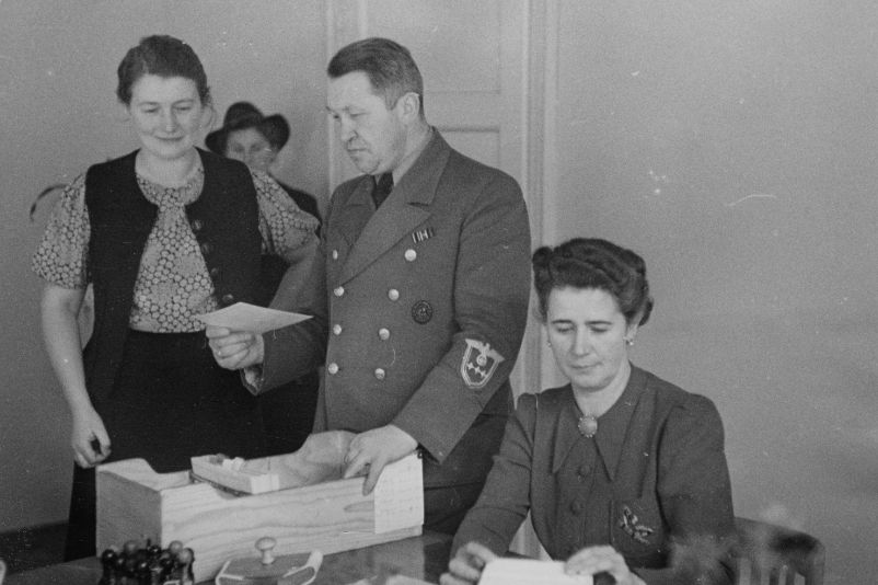 Das Bild zeigt den Innenraum eines Büros in einem Arbeitsamt in Polen. Ein Uniformierter Mitarbeiter wirft einen prüfenden Blick auf eine Kartei. Vor ihm am Tisch sitzt eine frau die ebenfalls Karteikarten bearbeitet. Links von ihm steht eine weitere Frau, die auf Ihn zu warten scheint.